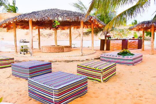 beach lounge area with thatched roof gazebos and colorful ottomans, luxury outdoor event setup on sand, tropical resort relaxation space with decorative string lights, square puffs , piassava kiosk 