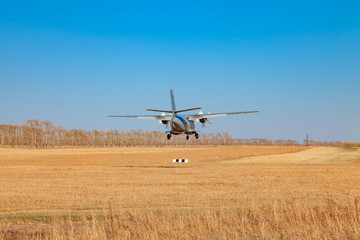 Fototapeta na wymiar A small aircraft for the transport of passengers and paratroopers lands in a field on a landing strip with grass under a blue sky above the trees on a clear cloudless day. Air patrol.