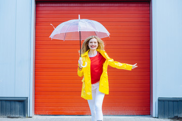 Inspired excited blond woman under umbrella in yellow raincoat, red t-shirt over red garage door background. Cool expression on face. Outdoor portrait..Weather forecast lifestyle concept