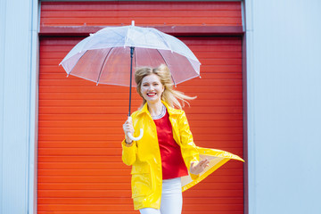 Inspired excited blond woman under umbrella in yellow raincoat, red t-shirt over red garage door background. Cool expression on face. Outdoor portrait..Weather forecast lifestyle concept