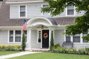 Neat and pretty shingled retro house with arched entryway and beautiful landscaping with colorful summer wreath on front door and American Flag attached to front column