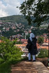 Fototapeta na wymiar Bosnia and Herzegovina: muslim woman from behind looking at the breathtaking skyline of Sarajevo, along the Miljacka River and surrounded by the Dinaric Alps , from the Zuta Tabija (Yellow Fortress) 
