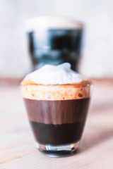 Double macchiato, espresso with milk froth in a small glass in front of a pint of stout with a froth on top in the background on a wooden table.