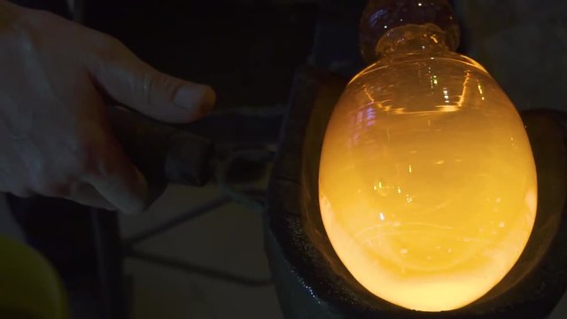 Glassblower in his workshop holding rod with liquid glass and turning it. Close-up.