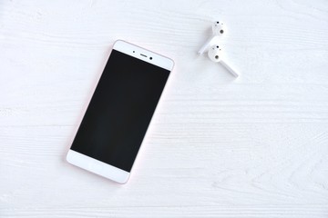 Smartphone with white wireless earphones with selective focus on white wooden background with empty space for text. Mobile phone and portable headphones on textured backdrop 