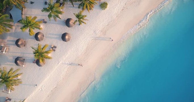 Aerial view of sea waves, umbrellas, palm trees and walking people on sandy beach at sunset. Summer in Zanzibar, Africa. Tropical landscape with parasols, sand, blue water. Top view from drone. Travel