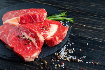 Three pieces of juicy raw beef on a stone cutting board on a black wooden table background.