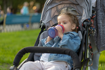 Little Caucasian girl dressed in a denim jacket drinks juice from her bottle while sitting in a baby carriage