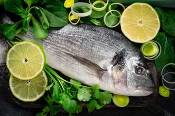 Fresh and raw sea bream fish with lemon and herbs on a black wooden background, top view and close...