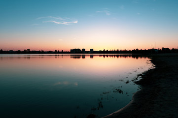 Sunset above the lake and city beach in summer with dark silhouettes of houses