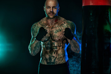 Sportsman with tattoos, man boxer fighting in gloves with boxing punching bag. Isolated on black...