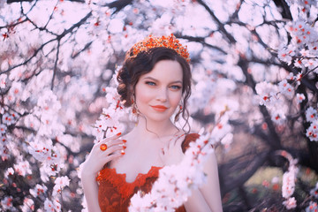 Fototapeta premium Queen of Spring walks in the blooming garden, a portrait photo of a pretty woman with dark hair and a golden crown on her head, the royal greatness, a red lady with bright blue eyes in sakura flowers