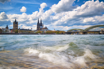 Kölner Dom, Groß St. Martin Kirche, Hohenzollernbrücke, Altstadt Köln, Rheinboulevard, Rheinufer, Wellen im Rhein, Blauer Himmel mit weißen Wolken.
