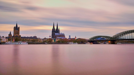 Fototapeta premium Sonnenuntergang in Köln am Rhein mit Orange Roten Farben am Himmel und Horizont. Blick auf Kölner Dom, Hohenzollernbrücke, Groß St, Martins Kirche und die Kölner Altstadt. Panoarama Hintergrund Poster