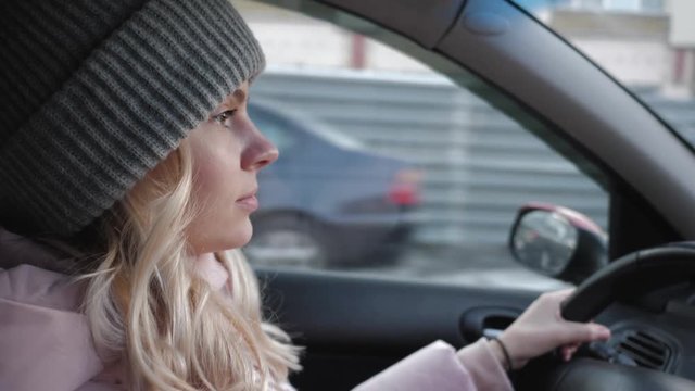 Young pretty blond woman with long curly hairstyle in grey sport knit hat, pink parka driving car through a city road traffic, rotating steering wheel, looking around, maneuver, give way to