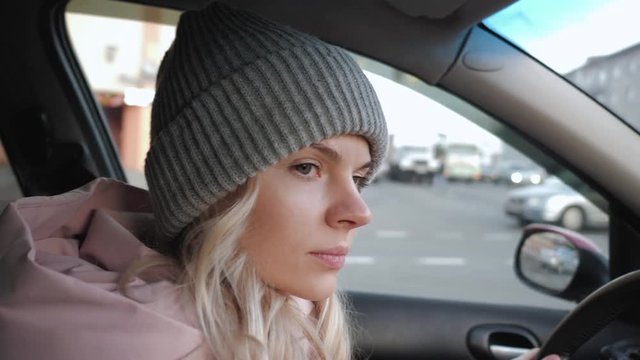 Young pretty blond woman with long curly hairstyle in grey sport knit hat, pink parka driving car through a city road traffic, rotating steering wheel, looking around, maneuver, give way to