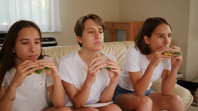 Triplets sisters teenager girls at home watch tv sit on a couch eating tasty sandwiches together