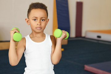 Cute boy athlete of African appearance doing physical exercises at gym after school, holding two green dumbbells, pumping arms. Dark skinned male child dressed in white tank top training indoors