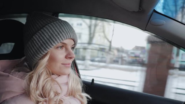 Young pretty blond woman with long curly hairstyle in grey sport knit hat, pink parka driving car through a city road traffic, rotating steering wheel, looking around, maneuver, give way to