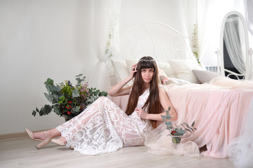 Photo of a young girl in the style of fine art. The girl in a white translucent dress sits on the floor next to the bed and looks into the camera. Girl in dress sits on laminate floor