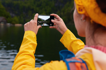 Naklejka premium Back view of unrecognizable female model makes photo on smartphone, captures beautiful view in screen, wears yellow raincoat, takes pictures for sharing impressions about journey. Focus on hands