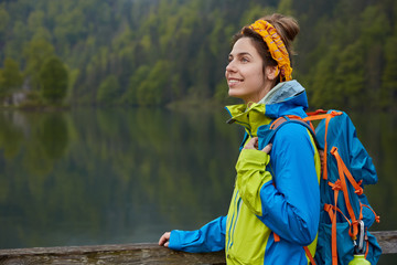 Outdoor view of happy active female wanders near lake and green forest, enjoys majestic landscape, stands sideways at camera on wooden bridge, focused into distance with dreamy facial expression