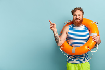 Happy redhead man with curly hair, relaxes at summer beach, poses with bright orange lifebuoy, points left, cares about safe journey, shows something on blue wall. Rescue and safety on water