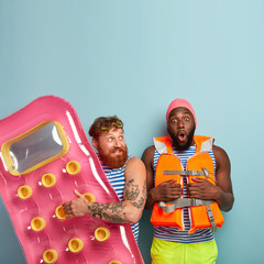 Funny bearded red haired man holds inflated mattress, shocked dark skinned man afraids of swimming,...