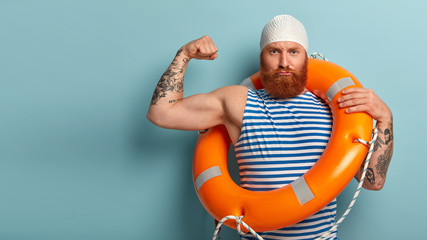 Self confident strong male lifeguard shows muscles, raises arm, has serious expression, carries safety ring, cares about water safety, helps swimmers who are in trouble, responds on aquatic emergency - obrazy, fototapety, plakaty