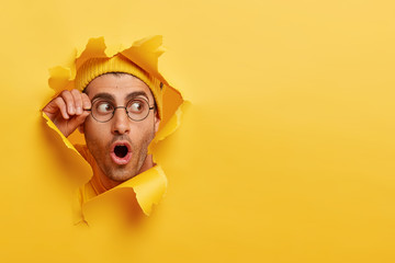 Astonished unshaven man gazes through hole in yellow paper, wears round spectacles, opens mouth...