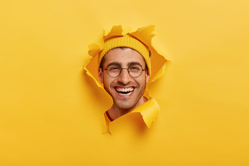 Headshot of positive unshaven young man smiles broadly, wears round optical glasses, yellow headgear, enjoys good time while making photos, looks through torn paper hole, expresses happiness