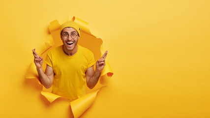 Hopeful excited man makes wish, crosses fingers, smiles joyfully, believes in good luck, stands at torn hole, wears yellow t shirt in one tone with background. Monochrome. Fullfilling dream.