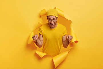 Studio shot of happy man with joyful facial expression, points down on floor, promots something, shows direction in bottom, demonstrates advert, recommends product, stands in torn hole of yellow wall