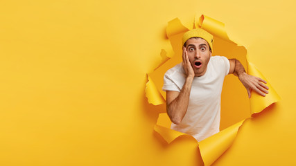 Impressed young man touches cheek, has stupefied facial expressions, drops jaw and stares at camera, looks out of hole in paper, cuts through yellow wall with empty space for your information