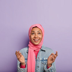 Religious cheerful woman has Islamic views, raises palms, prays and looks hopefully upwards, wrapped in pink hijab, isolated over purple background with blank space for your slogan, holds hands up