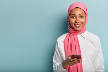 Pleased Muslim woman uses cell phone to socialize, gives reply in online chat, posts something in social networks, wears pink veil and white shirt, isolated over blue background, blank space aside