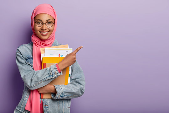 Satisified Muslim college student with notepad and papers, wears round spectacles and denim jacket, points away on blank space, isolated over purple background. Religious views and studying concept