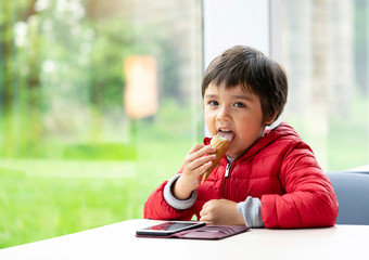 Happy child licking an ice cream, kid boy sitting alone watching cartoon on mobile phone and eating an ice cream, Relaxing preschool boy sitting at the table in a cafe  with blurry natural background.