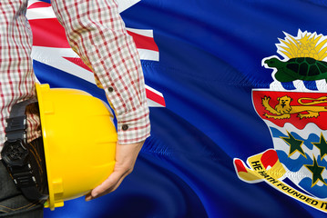 Engineer is holding yellow safety helmet with waving Cayman Islands flag background. Construction...