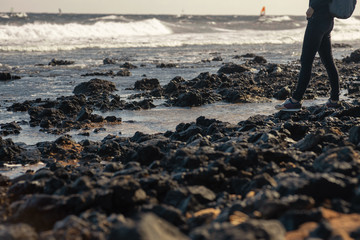 a walk along the coast of volcanic stones, a girl in jigs with a backpack stands on the rocky shore of the ocean, in the background you can see wind surfers. No face.