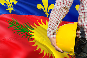 Engineer is holding yellow safety helmet with waving Guadeloupe flag background. Construction and building concept.