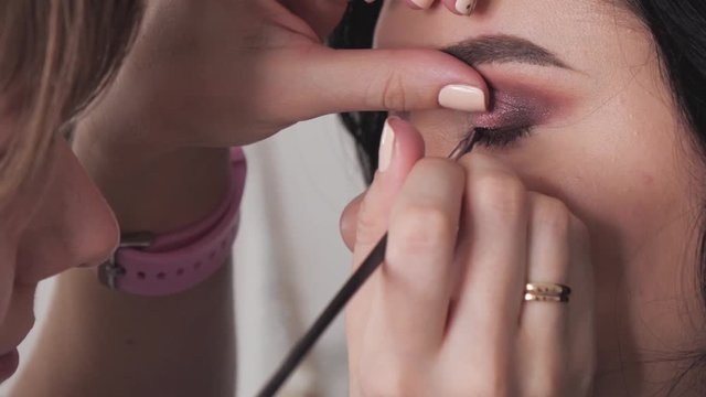 Closeup shot of cheerful young female makeup artist enjoying working with a beautiful young brown haired model, applying fresh trendy makeup on her eyes with long eyelashes, perfect brows using pencil