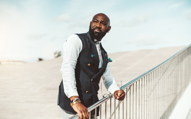 Portrait of a serious noble bald bearded African man in an elegant vest standing outdoors with the...