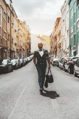 Vertical shot of an elegant adult bald black guy with a beard and in spectacles&nbsp;and fashion vest, with a leather bag in his hand descending down the narrow Lisbon street with plenty of cars on sides