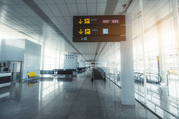 Wide-angle view of a long empty hall of a modern airport terminal with a bright glass facade on the right, the signpost with arrows and information, the travelator, gates, and rows of plastic seats - obrazy, fototapety, plakaty