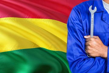 Bolivian Mechanic in blue uniform is holding wrench against waving Bolivia flag background. Crossed arms technician.