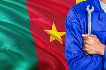 Cameroonian Mechanic in blue uniform is holding wrench against waving Cameroon flag background. Crossed arms technician.