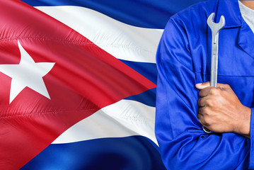 Cuban Mechanic in blue uniform is holding wrench against waving Cuba flag background. Crossed arms technician.