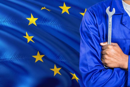 Mechanic in blue uniform is holding wrench against waving European Union flag background. Crossed arms technician.