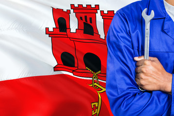 Mechanic in blue uniform is holding wrench against waving Gibraltar flag background. Crossed arms...